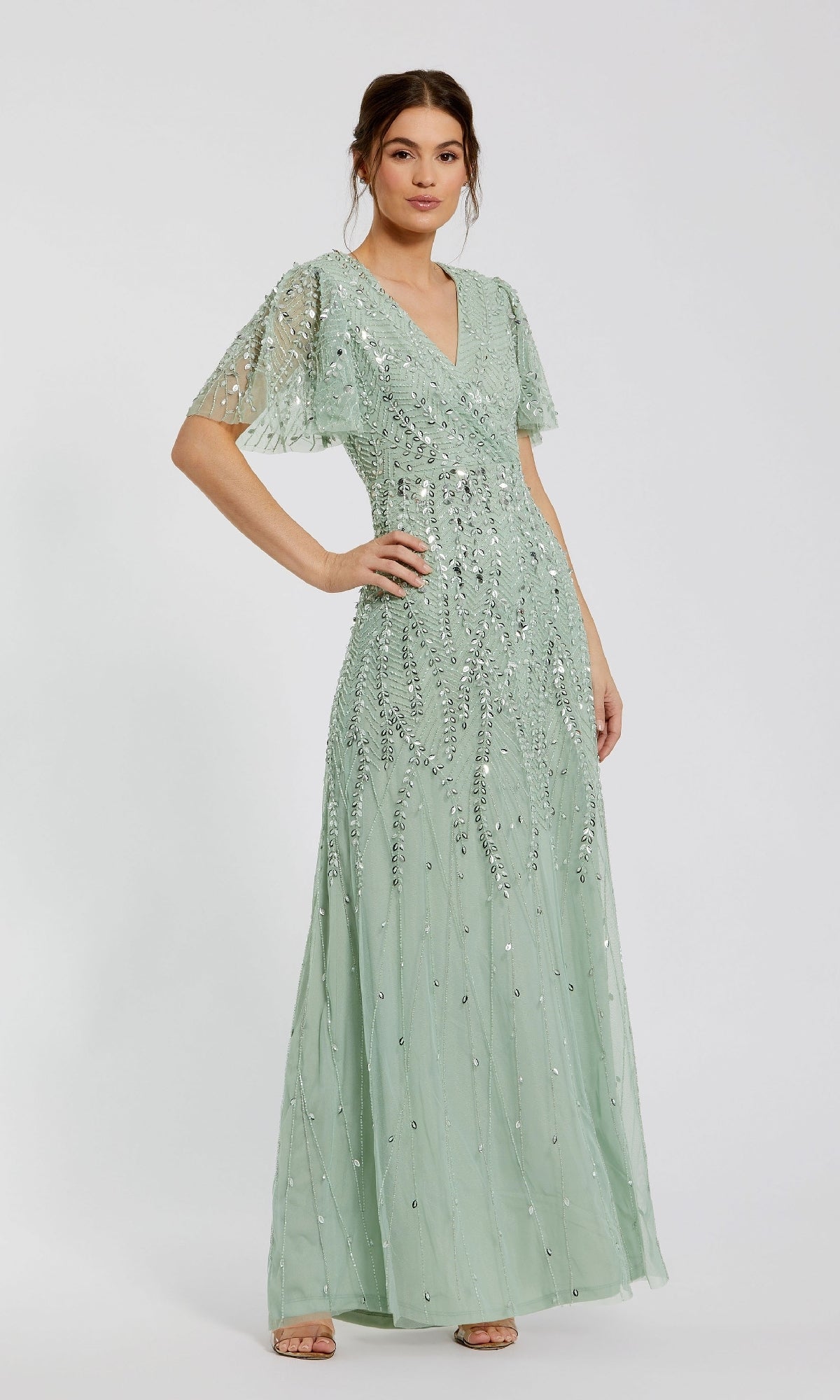 A woman models the Mac Duggal 35109 Flutter-Sleeve Long Formal Dress, a floor-length light green gown with short flutter sleeves and silver sequin accents, standing confidently with one hand on her hip against a plain background.